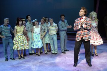 Lavalla laulaa ja hymyilee joukko 1950-luvun värikkäisiin pukuihin pukeutuneita esiintyjiä, joista yksi ruututakkinen mies seisoo edessä ja näyttää johtavan. Tausta on sininen, ja tunnelma on vilkas ja iloinen.