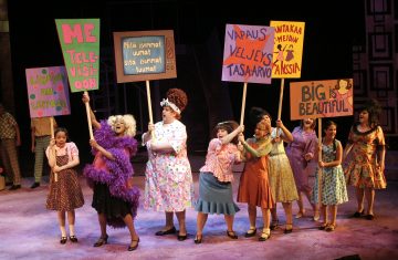 Lavalla on joukko värikkäisiin asuihin pukeutuneita ihmisiä, jotka pitävät mielenosoituskylttejä, joissa on erilaisia iskulauseita. He näyttävät esittävän vilkasta musikaalia tai teatteriesitystä.