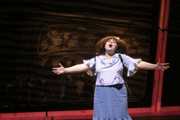 Siniseen pilkkupuseroon ja siniseen hameeseen pukeutunut esiintyjä laulaa intohimoisesti lavalla kädet ojennettuina ja seisoo taustan edessä, jossa on vintage-tyylinen taideteos autossa istuvista ihmisistä.