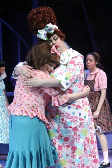 Kaksi värikkäisiin pukuihin pukeutunutta naista halaa lohduttavan halauksen lavalla, toisella on suuri rusetti ja kiharat punaiset hiukset. Toinen ruutupukuinen nainen seisoo taustalla ja tarkkailee kohtausta. Näyttää siltä, että kyseessä on teatteriesitys.