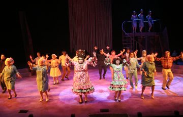 Vilkas lavaesitys, jossa suuri joukko värikkäisiin pukuihin pukeutuneita esiintyjiä tanssii kirkkaiden valojen alla; kolme esiintyjää, joilla on röyhelöisiä mekkoja, seisovat keskellä, kun muut tanssivat heidän ympärillään. Kolme laulajaa seisoo yläpuolella korokkeella.