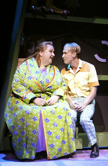 Kaksi näyttelijää istuu näyttämöllä tiiviisti vierekkäin. Vasemmalla olevalla henkilöllä on kirkkaanvihreä kukkatakki, kun taas oikealla olevalla on keltainen paita ja ruudulliset housut. Molemmat näyttävät näyttämövalaistuksessa keskustelevan keskenään.