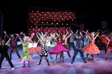 Suuri joukko värikkäisiin, retrotyylisiin pukuihin pukeutuneita esiintyjiä tanssii energisesti lavalla kädet kohotettuna eloisan, kuviollisen taustan ja näyttämövalojen edessä.