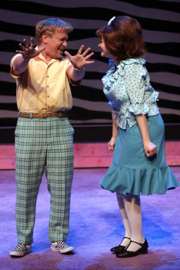 Keltaiseen paitaan ja ruudullisiin housuihin pukeutunut mies osoittaa innostuneesti hymyilevälle naiselle, jolla on sininen pilkkupusero ja teerihame, kun he seisovat lavalla, molemmat pukeutuneina eloisiin, retrotyylisiin pukuihin.