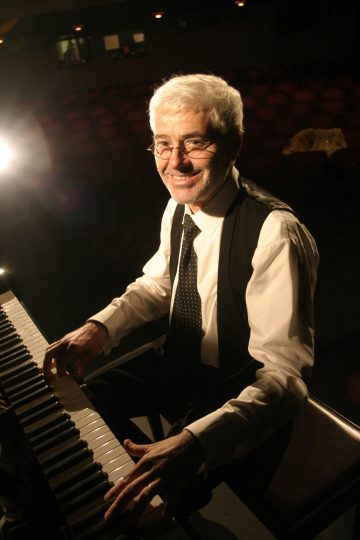 Vanhempi mies, jolla on valkoiset hiukset ja silmälasit ja jolla on valkoinen paita, musta liivi ja solmio, hymyilee soittaessaan pianoa hämärässä auditoriossa näyttämövalaistuksen alla.