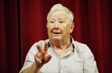 Vanhempi mies, jolla on valkoiset hiukset ja vaalea paita, osoittaa kädellään kolme sormea ylöspäin. Hän seisoo runsaan punaisen verhon edessä ja katsoo hieman ylöspäin ilmeikäs ilme kasvoillaan.