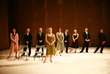 Yhdeksän viralliseen ja puoliviralliseen asuun pukeutunutta ihmistä seisoo rivissä näyttämöllä, jossa on ruskea tausta; kaksi tyhjää tuolia on sijoitettu heidän eteensä, ja lämmin näyttämövalaistus korostaa kohtausta.