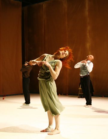 Lavalla esiintyy kolme nykytanssijaa; edessä oleva tanssija, jolla on punaiset hiukset ja vihreä mekko, kumartuu dramaattisesti taaksepäin, kun taas kaksi tanssijaa taustalla poseeraa kädet kaulan lähellä. Lava on lämpimästi valaistu.