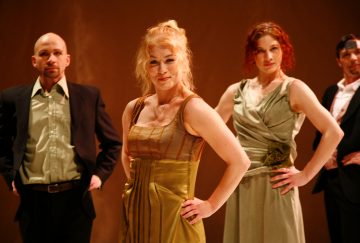 Neljä ihmistä seisoo itsevarmasti kädet lanteilla, pukeutuneina juhlavaatteisiin. Kaksi kullanvihreään mekkoon pukeutunutta naista on etualalla, ja taustalla on kaksi pukuun pukeutunutta miestä, jotka kaikki ovat kasvot menosuuntaan lämmintä, ruskehtavaa taustaa vasten.