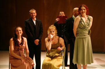 Lavalla on kuusi muodollisesti pukeutunutta aikuista. Edessä on kolme pukuun pukeutunutta naista; yksi istuu ja pyyhkii kasvonsa nenäliinalla, kaksi seisoo ja hymyilee. Takana seisoo kolme pukumiestä, joista yhdellä on kirja kädessään ja kaksi muuta hymyilee.