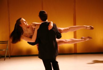 Pukumies nostaa ja pyörittää hymyilevää, lyhyeen mekkoon pukeutunutta naista tanssiesityksessä, jonka vartalo on ojennettu vaakasuoraan. Tausta on lämpimästi valaistu ja vasemmalla näkyy tyhjä tuoli.