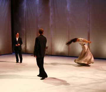 Kolme ihmistä on lavalla, jossa on pehmeä valaistus; kaksi pukumiestä seisoo katsomassa, kun virtaavaan mekkoon pukeutunut nainen pyörii tai tanssii tarmokkaasti, ja hänen pitkät hiuksensa ja mekkonsa liikkuvat dramaattisesti.