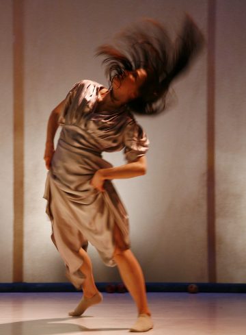 Löysään, vaaleaan mekkoon pukeutunut henkilö tekee dynaamisen tanssiliikkeen ja kääntää dramaattisesti pitkiä tummia hiuksiaan seisoessaan yhdellä jalalla pehmeästi valaistua taustaa vasten.