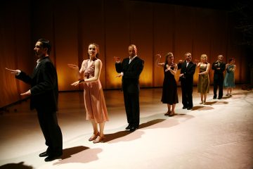 Kahdeksan viralliseen ja puoliviralliseen asuun pukeutunutta ihmistä seisoo lavalla rivissä kädet taivutettuina ja kämmenet ylöspäin ja tekee synkronoituja liikkeitä lämpimästi valaistua taustaa vasten.