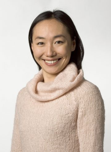 Nainen, jolla on suorat tummat hiukset ja päällään vaaleanpunainen neulepusero, jossa on cowl-kaulus, hymyilee kameralle valkoista taustaa vasten.