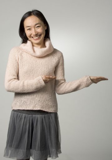 Hymyilevä nainen, jolla on pitkät tummat hiukset, vaaleanpunainen pusero ja harmaa hame, seisoo yksinkertaista taustaa vasten ja osoittaa molemmilla käsillään sivulleen kuin esittelisi jotain.