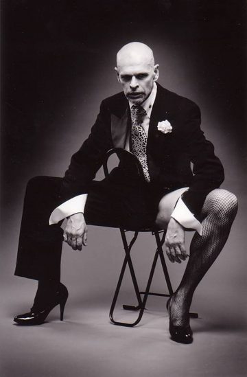 Kalju mies, jolla on smokkitakki, pukupaita ja solmio, istuu selinmakuulla tuolilla jalat levällään, jalassaan verkkosukat ja korkokengät. Kuva on mustavalkoinen, ja hänellä on vakava ilme.