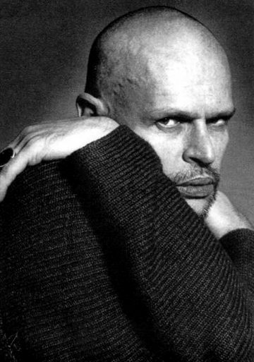 Kalju henkilö, jolla on vakava ilme, lepuuttaa kätensä pinnalla, ja hänellä on yllään tumma, kuvioitu villapaita. Valaistus on dramaattinen ja korostaa hänen kasvojensa ja päänsä ääriviivoja. Tausta on yksinkertainen ja tumma.