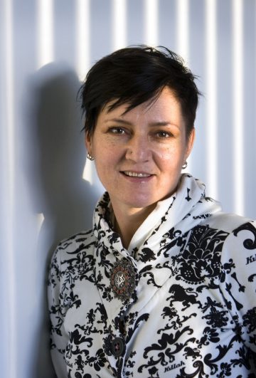 Lyhyet tummat hiukset omaava henkilö, jolla on yllään valko-mustakuvioinen takki, jossa on koristeellinen rintakoru, seisoo seinää vasten, jossa on pystysuuntaisia varjoja, ja hymyilee kameralle.