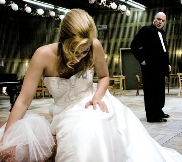 Valkoiseen kaapuun pukeutunut nainen istuu vaaleanpunaisella sohvalla eteenpäin nojautuneena pää alaspäin. Taustalla seisoo mustaan smokkiin pukeutunut mies, joka katsoo häntä. Ympäristö vaikuttaa muodolliselta, modernilta huoneelta, jossa on puupanelointi.