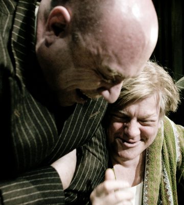 Kaksi ihmistä, joista toisella on kalju ja raidallinen takki ja toisella vaaleat hiukset ja vihreä pyyhe, näyttävät lähikuvassa nauravan tai jakavan tunteikkaan hetken yhdessä.