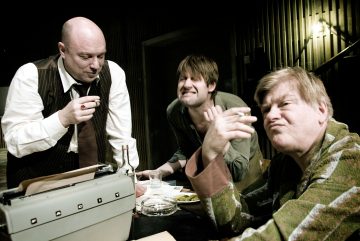 Kolme miestä istuu pöydän ääressä juomien, papereiden ja kirjoituskoneen kanssa. Yksi kaapuun pukeutunut mies pitelee savuketta ja tekee ilmeen, toinen nojaa eteenpäin virnistäen, ja kolmas katsoo heitä mietteliäästi. Asetelma vaikuttaa teatraaliselta.