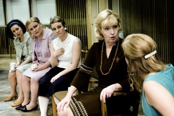 Viisi naista istuu rivissä puutuoleilla keskustellen. Useimmilla heistä on mekko, ja heillä on 1960-luvun tyyliset hiukset ja asusteet. Ruskeapukuinen nainen näyttää puhuvan, kun taas muut kuuntelevat tarkkaavaisesti.