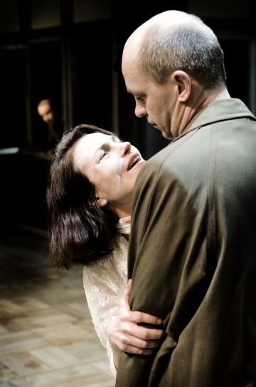 Tummahiuksinen nainen takertuu tunteella ruskeapukuiseen kaljuun mieheen ja katsoo häntä ahdistuneena. Kohtaus on sisätiloissa, ja taustalla on toinen hahmo, joka on hämärtynyt.