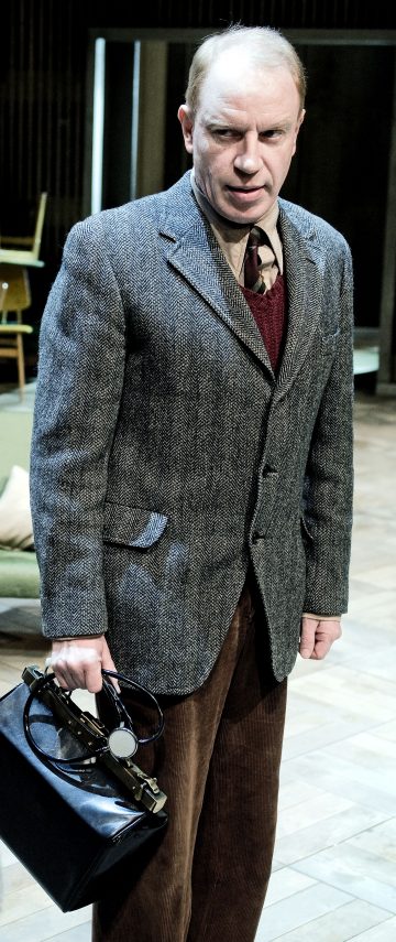 Vakavan näköinen mies, jolla on harmaa bleiseri, punainen pusero ja ruskeat korduroy-housut, seisoo sisätiloissa pitäen kädessään mustaa laukkua ja ilmeisesti lääkinnällisiä välineitä. Taustalla näkyy puulattioita ja huonekaluja.