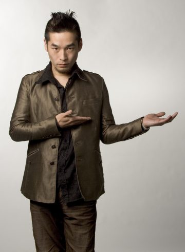 Mies, jolla on lyhyet, muotoillut tummat hiukset, ruskea puku ja musta paita, seisoo tavallista taustaa vasten ja osoittaa toisella kädellä avoimeen ja toisella kädellä kohti.