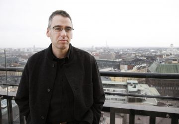 Silmälasipäinen ja tummatakkinen mies seisoo kattoparvekkeella, jolta avautuu näkymä kaupunkimaisemaan pilvisenä päivänä. Taustalla näkyy rakennuksia ja kaukainen kupoli.