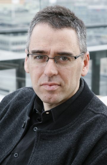 Lyhyet tummat hiukset, silmälasit, tumma takki ja paita, mies istuu ulkona ja katsoo suoraan kameraan neutraali ilme kasvoillaan; taustalla sumea kaupunkikuva.