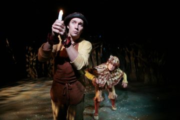 Maalaispukuinen henkilö pitelee sytytettyä kynttilää ja katsoo huolestuneena ylöspäin, kun toinen eläimeksi pukeutunut henkilö kyyristelee heidän takanaan hämärästi valaistulla näyttämöllä.