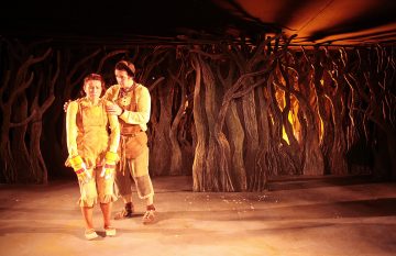 Kaksi näyttelijää seisoo näyttämöllä, jonka taustalla on kierrettyjä puun oksia. Toinen näyttelijä lohduttaa toista, joka näyttää ahdistuneelta. Molemmilla on haalaria muistuttavat puvut, joita valaisee lämmin, oranssi valaistus.