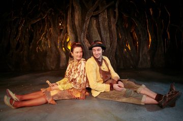 Kaksi näyttelijää värikkäissä satupuvuissa istuu lavalla selkä toisensa vasten ja hymyilee. Heidän takanaan on tumma, mutkitteleva, kierrettyjä puun juuria muistuttava tausta, jonka läpi paistaa lämmin valo.