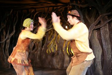 Kaksi maalaismaisiin, maanläheisiin pukuihin pukeutunutta ihmistä taputtaa leikkisästi käsiä yhteen lavalla, jonka taustalla on kieroutuneita, puiden kaltaisia rakenteita, jotka viittaavat omituiseen tai satumaiseen ympäristöön.