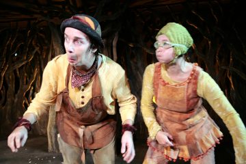 Kaksi näyttelijää, joilla on omituiset, maansävyiset puvut ja hatut, tekevät lavalla hauskoja, liioiteltuja ilmeitä, ja taustalla on vääntyneitä puun juuria tai oksia muistuttava tausta.