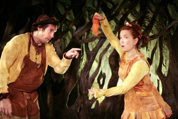 Kaksi keltaisiin ja ruskeisiin pukuihin pukeutunutta näyttelijää esiintyy kierrettyjen puunoksain edessä; toinen osoittaa ja toinen pitää oranssia kangasta ylhäällä, ja molemmilla on ilmeikkäät kasvot ja eleet.