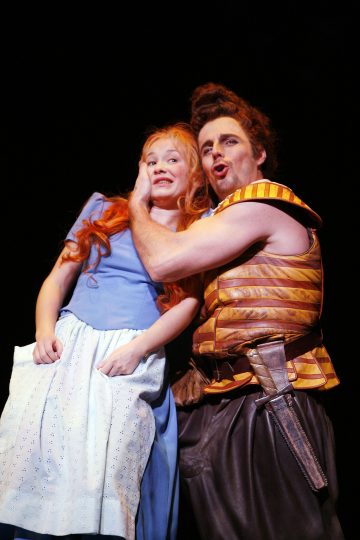 Siniseen mekkoon pukeutunut nuori tyttö näyttää levottomalta, kun raidalliseen keskiaikaiseen liiviin pukeutunut mies pitelee hänen kasvojaan ja laulaa dramaattisesti lavalla mustaa taustaa vasten.