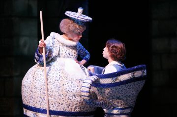 Kaksi esiintyjää, jotka ovat pukeutuneet hienostuneisiin sinivalkoisiin teekannu- ja teekuppiasuihin ja jotka todennäköisesti esittävät Kaunottaren ja hirviön hahmoja, kohtaavat toisensa lavalla tummaa teatteritaustaa vasten.