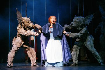 Sinivalkoiseen mekkoon pukeutunut nainen näyttää ahdistuneelta, kun kaksi kivenkaltaisiin gargoyle-asuihin pukeutunutta ihmistä tarttuu hänen käsiinsä dramaattisella, hämärästi valaistulla näyttämöllä.