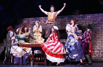 Liiviin pukeutunut mies seisoo riemukkaasti pöydällä ja taivuttaa käsiään, ja häntä ympäröivät vanhanajan värikkäisiin mekkoihin ja hattuihin pukeutuneet hurraavat naiset näyttämöllä teatteriesityksessä, jonka taustana on tiiliseinä.