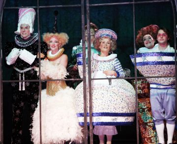 Ryhmä näyttelijöitä seisoo ikkunakehyksen takana hienostuneissa, värikkäissä puvuissa, pukeutuneina oudoiksi hahmoiksi, kuten teekannuksi, teekupiksi ja höyhenpeitteiseksi hahmoksi, jotka ovat todennäköisesti peräisin teatterituotannosta.