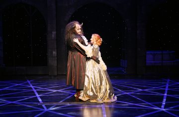 Hirviöasuun pukeutunut henkilö ja kultaiseen pukuun pukeutunut henkilö tanssivat yhdessä lavalla, jonka taustalla on tumma tähtitausta, joka muistuttaa kohtausta Kaunottaresta ja hirviöstä.