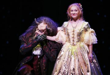 Henkilö, joka on pukeutunut taidokkaaseen Beast-pukuun, polvistuu ja pitelee hymyilevän naisen kättä, joka on pukeutunut kultaiseen, koristeelliseen pukuun, ja molemmat seisovat pimeällä, teatterin lavalla.