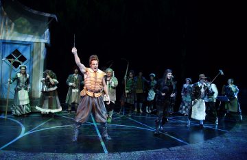 Ryhmä värikkäisiin keskiaikaisiin pukuihin pukeutuneita näyttelijöitä esiintyy lavalla, ja yksi näyttelijä edessä nostaa sauvaa dramaattisesti, kun taas muut seisovat taustalla pitelemässä rekvisiittaa ja katsomassa tarkkaavaisesti.