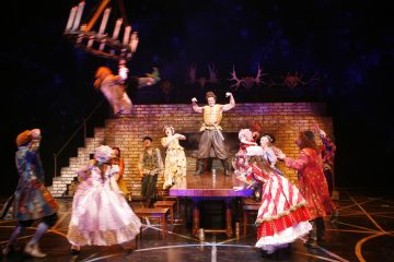 Teatterilavalla nähdään mies, joka jäntevöityy pöydällä, kun värikkäisiin pukuihin pukeutuneet ihmiset reagoivat dramaattisesti, ja yksi henkilö heiluu heidän yläpuolellaan olevasta kattokruunusta. Taustalla on portaikko ja sarvilla koristeltu tiiliseinä.