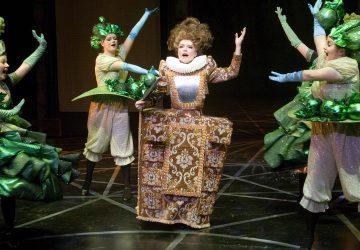 Keskellä näyttämöä seisoo koristeelliseen Elisabetilaistyyliseen pukuun pukeutunut esiintyjä, jota ympäröi neljä vihreisiin kiiltäviin asuihin ja hanskoihin pukeutunutta tanssijaa, jotka kaikki poseeraavat eloisasti teatteriesityksen aikana.