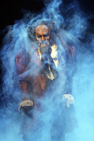 Kaunottaresta ja hirviöstä tuttuun Hirviöön pukeutunut henkilö seisoo dramaattisessa sinisessä sumussa, päällään riekaleinen ruskeankeltainen takki, valkoinen paita ja käsineet, sarvet ja hirviömäinen meikki.