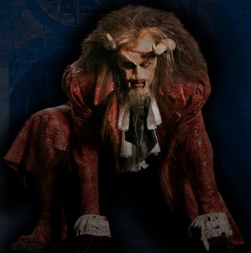 Henkilö, joka on pukeutunut hienostuneeseen petopukuun, jossa on sarvet, torahampaat ja villi tukka, ja jolla on yllään punainen, kuvioitu takki ja pitsiset hihansuut, kyykistelee nelinkontin tummaa, varjoista taustaa vasten.